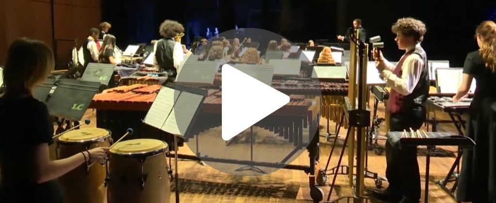 Captura de pantalla de una grabación de vídeo de un concierto navideño, con el fotógrafo disparando desde la parte trasera del escenario hacia el público, mostrando a los estudiantes tocando en una orquesta.