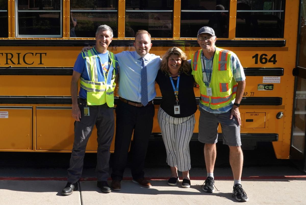 From left, a man in a reflective vest, a man in a dress shirt and tie, a woman in business casual wear, and a male bus driver stand beside each other and smile at the camera. In the background is a Poudre School District bus.
