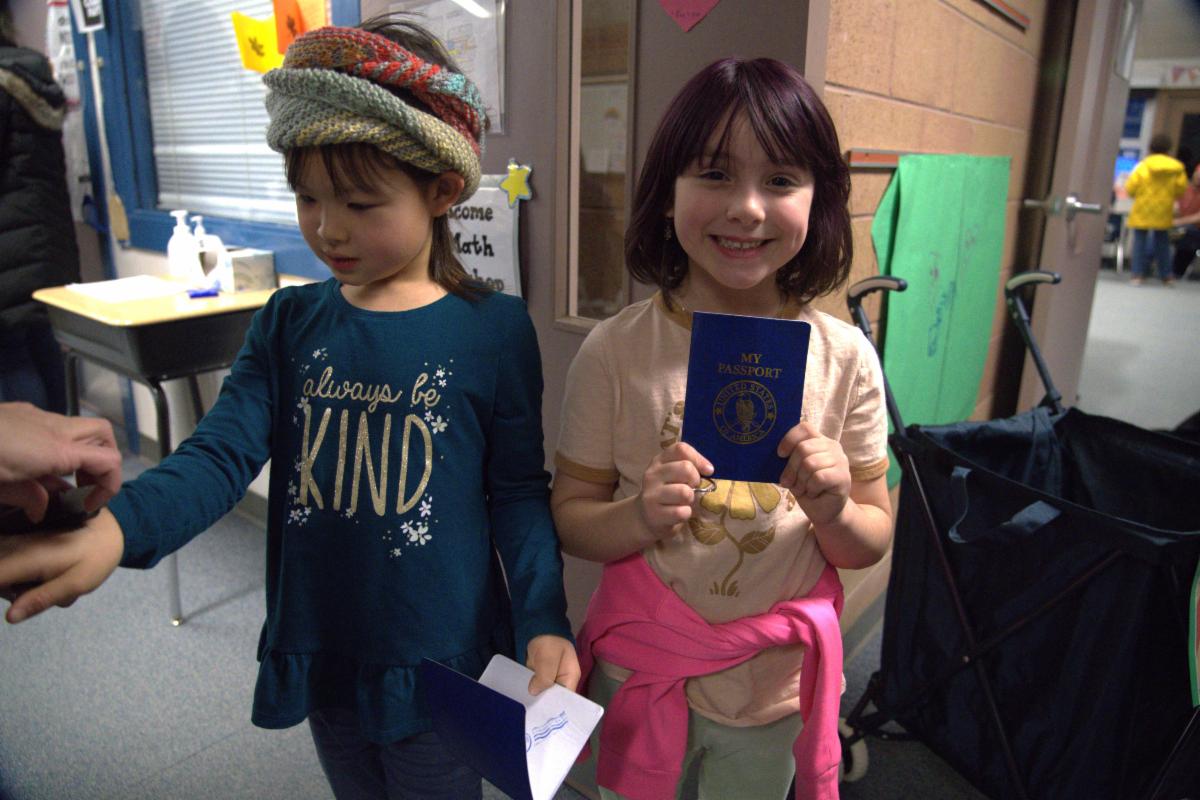 Two elementary school girls stand next to each other. One is looking at her hand while she gets a rubber stamp on it, and the other is smiling at the camera and holding a blue passport in front of her.