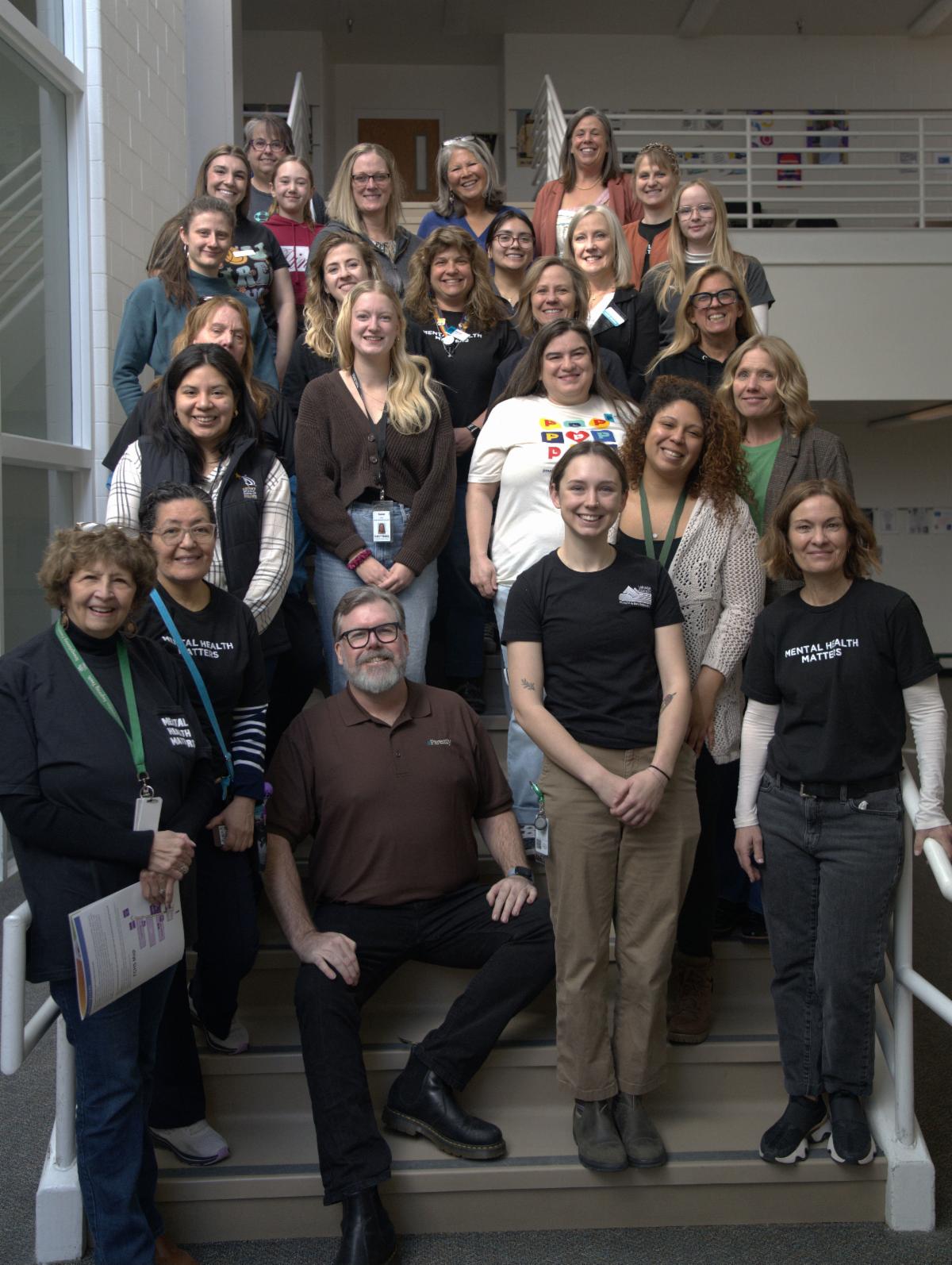 26 adults line up next to each on a staircase at Fort Collins High School for a photo. Some wear shirts that say, "Mental Health Matters."