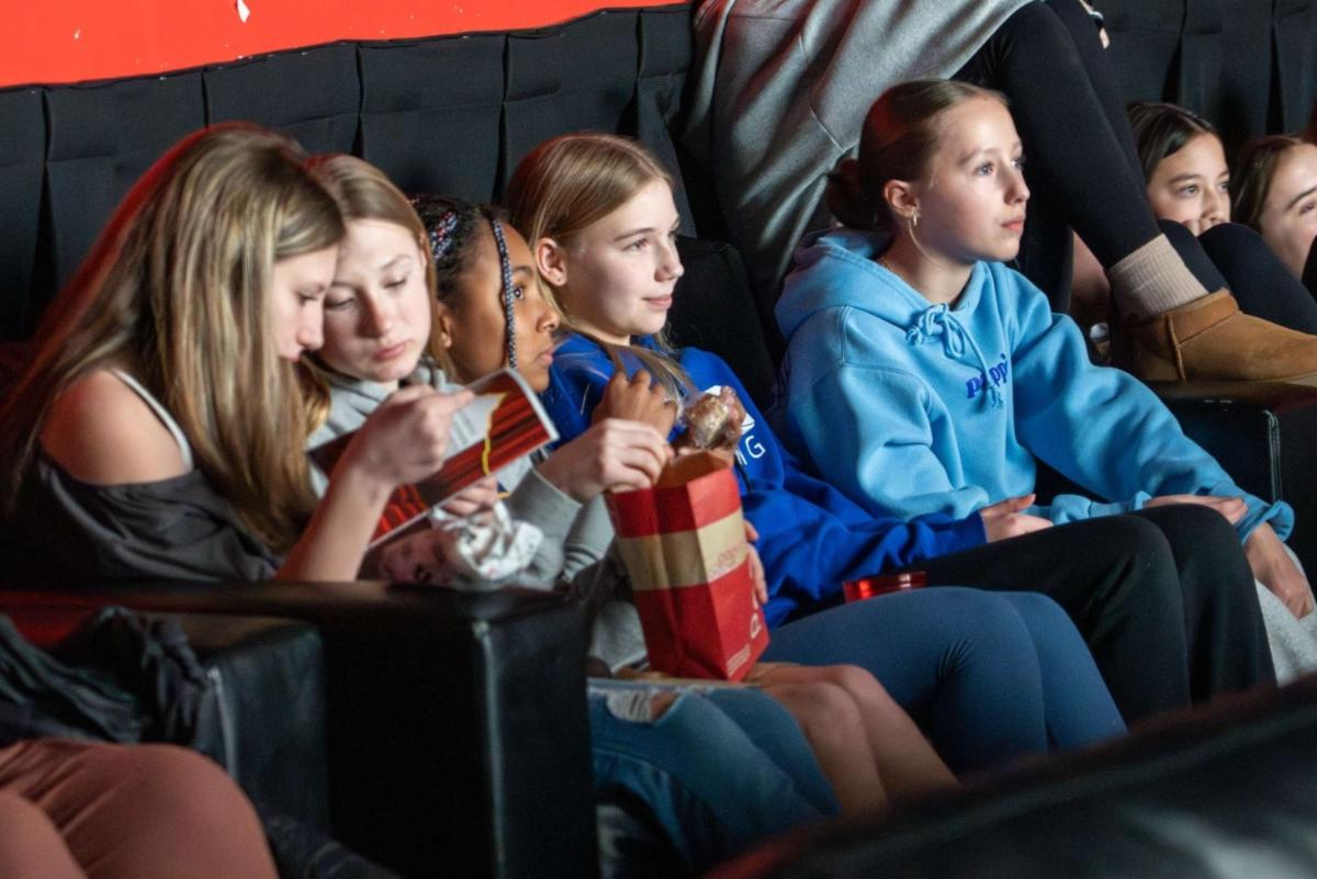 A group of middle school girls sit next to each other at a movie theater and watch the big screen.