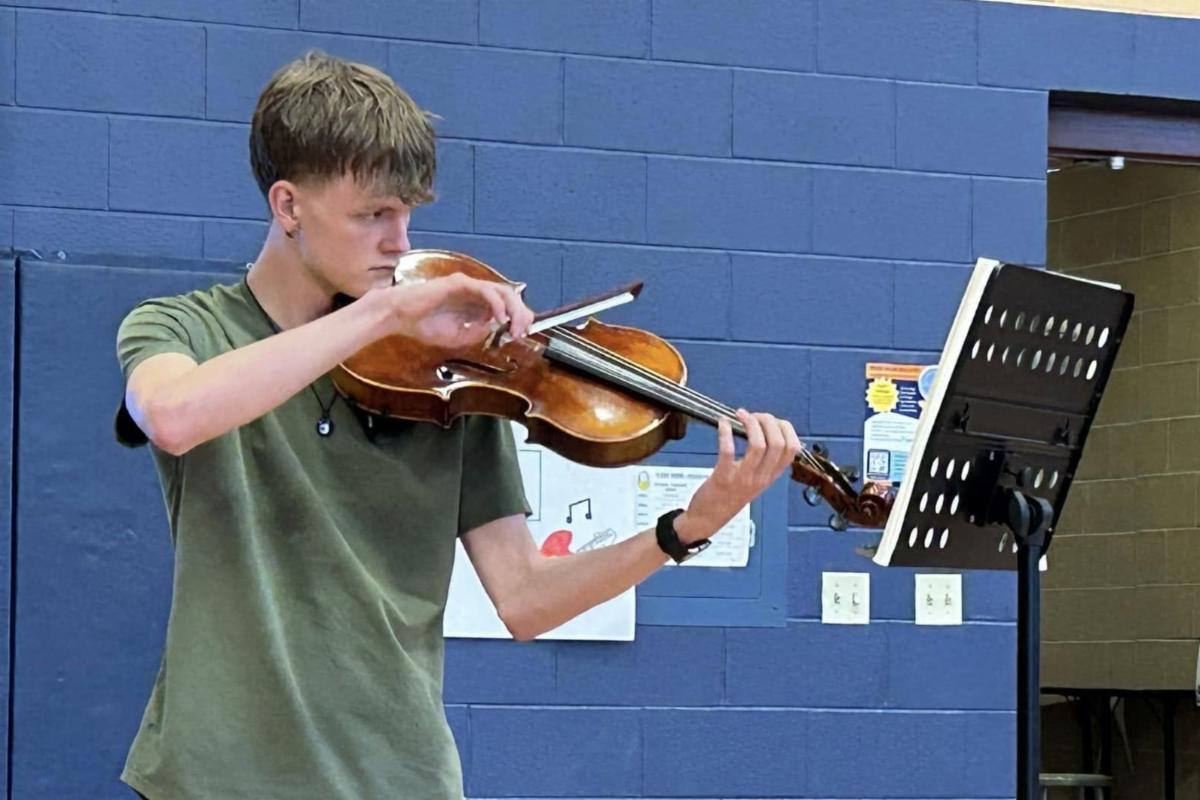 A high school boy plays a viola while standing in front of a stand holding sheet music.