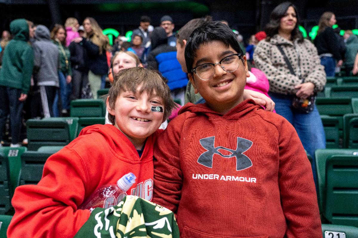 Two boys in red sweatshirts stand next to each other in the stands at Moby Arena and smile at the camera. One of the boys has a patch under his eye with the CSU Rams logo on it.