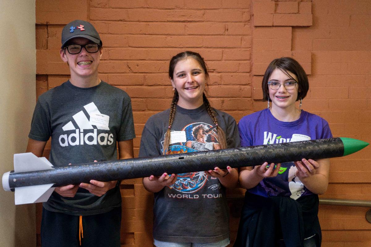 Tres estudiantes sostienen un modelo de cohete mientras están uno al lado del otro en un pasillo.