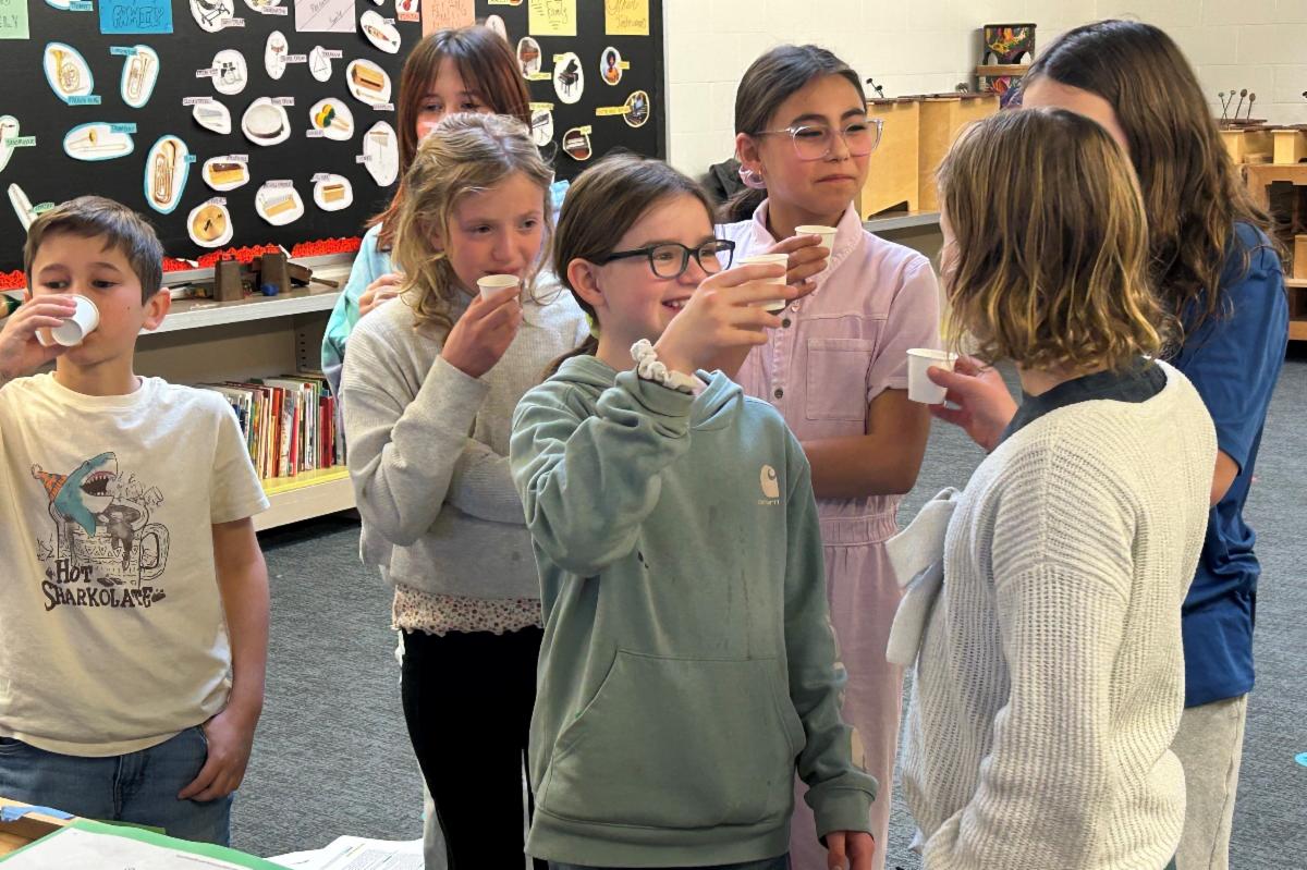 Seven upper elementary school students stand together in a classroom and cheers small paper cups filled with juice.