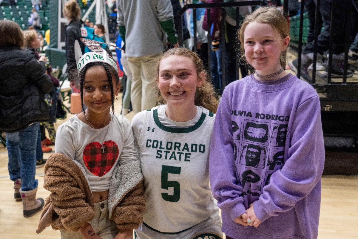Two girls stand on either side of a CSU women's basketball player wearing a jersey that says "Colorado State" and the number five as they pose for a photo.