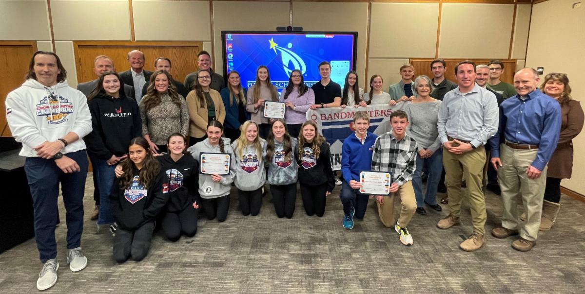 Several students and adults gather for a group photo in the school board meeting room. Some hold certificates or a banner for the state marching band championship.