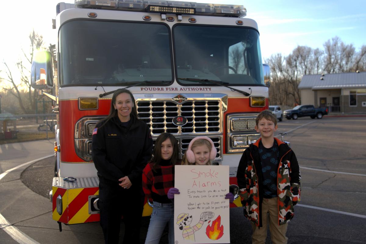 Una mujer con uniforme de bombero de la PFA junto a tres estudiantes de la PGA que ganaron un concurso de carteles. Los cuatro están parados frente a un camión de bomberos en el estacionamiento de la PGA. La estudiante parada en el medio sostiene su cartel, que tiene un mensaje de prevención de incendios y un dibujo de un dálmata con un sombrero contra incendios.