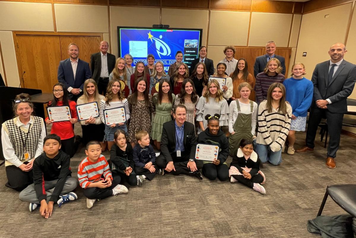 Treinta y cinco niños y adultos, de pie o sentados en cuatro filas, sonríen a la cámara en la sala de reuniones de la junta escolar.