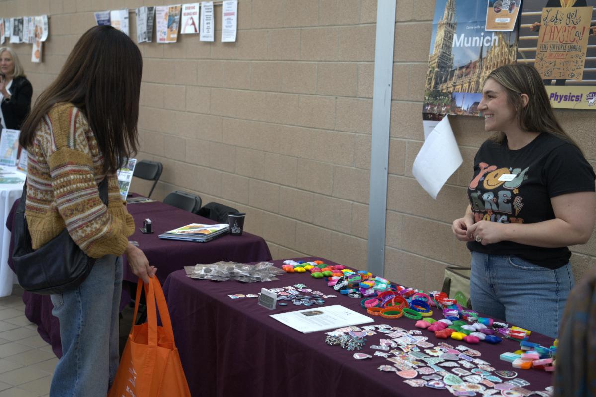 Two women stand on either side of a booth table at a resource fair along a hallway in Fort Collins High School. On top of the table are several swag items and information about mental health resources.
