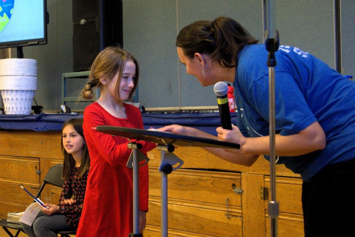 An elementary school girl in a red dress shakes the hand of her assistant principal while accepting a certificate in front of a stage in a gym.