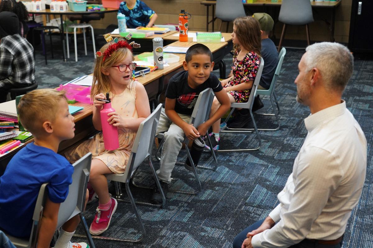 Four elementary school students seated side-by-side at desks in a classroom turn around to talk with a school board member, who is kneeling to speak at their level.