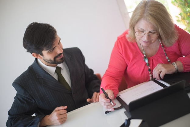 A senior woman signing paperwork with a young professional assisting her at a desk. (Stock Photo/Kampus Productions)