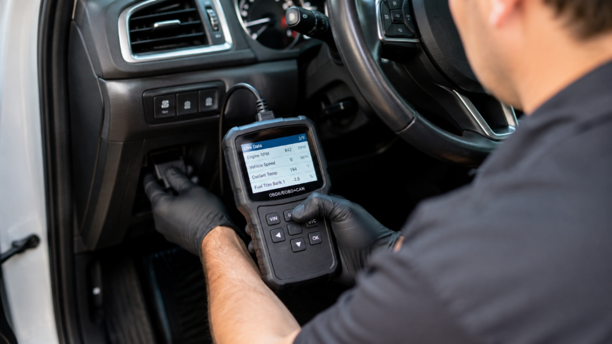 Automotive technician connecting an OBD-II diagnostic scanner under a car dashboard, with live engine data displayed on the scanner screen.
