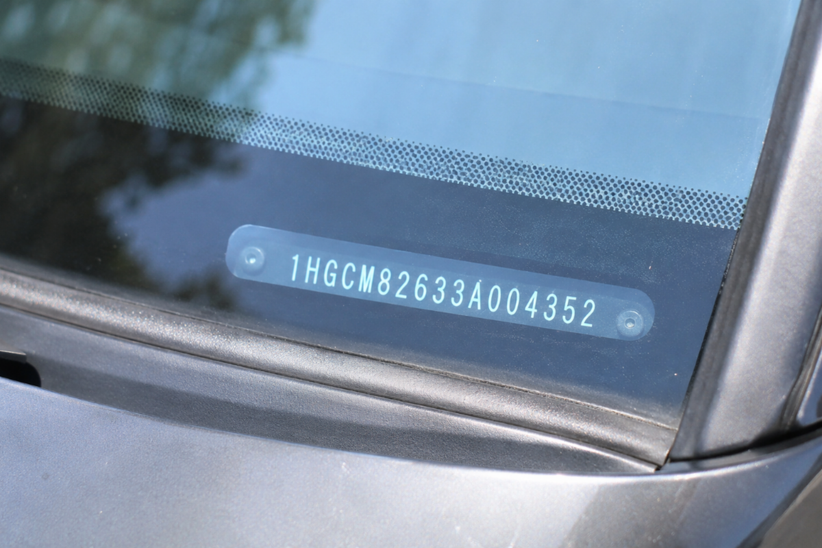 Close-up photo of a car dashboard viewed through the windshield, showing a VIN number plate in natural daylight with soft reflections on the glass and a shallow depth of field.