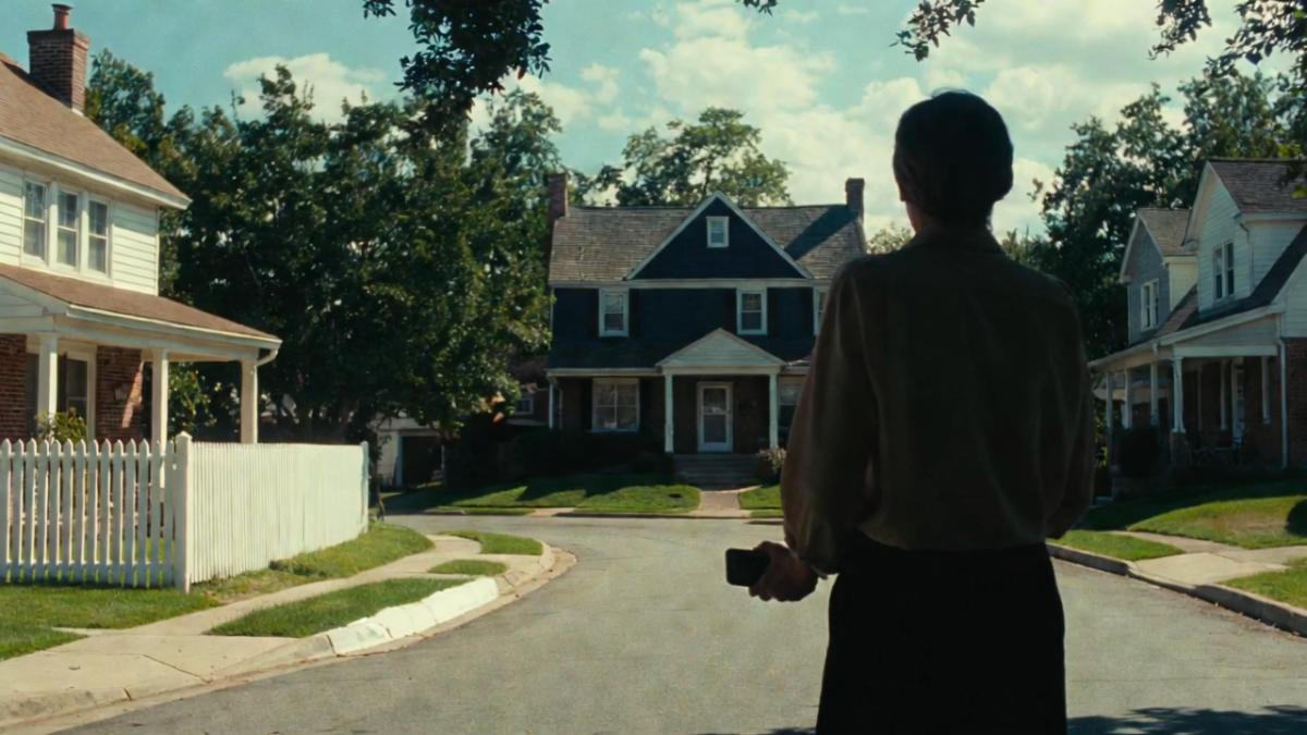 A middle-aged person with short hair, wearing a long-sleeved shirt and long skirt/pants, stands on a peaceful residential street in an American suburb. They are viewed from behind, holding a smartphone in their right hand. In the background, a classic two-story house with a dark roof, brick chimney, white siding, and front porch is visible under warm natural daylight. The street is lined with green lawns, mature trees, and other similar homes, creating a calm, sunny neighborhood atmosphere.