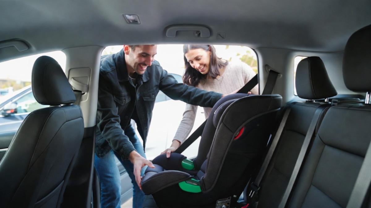 A smiling couple installing a black and gray infant car seat in the backseat of their car. The man in a dark jacket and the woman in a light sweater lean in through the open rear door, happily adjusting the straps and base together on a sunny day.