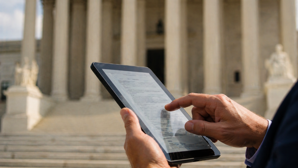 Close-up of hands scrolling through legal documents on a tablet, with a blurred courthouse building in the background under natural daylight, creating a clean, editorial-style scene.