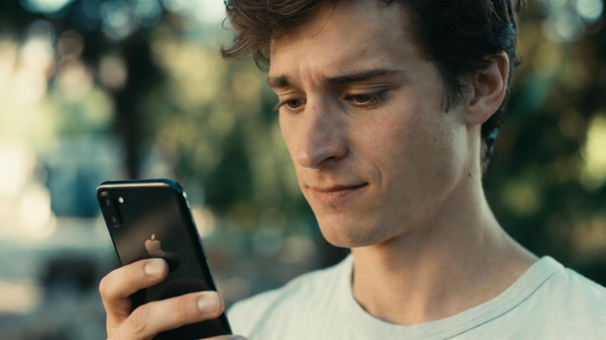 A young Caucasian man with curly brown hair and light stubble, wearing a light gray t-shirt, stands outdoors in soft evening light looking down thoughtfully at a black iPhone he holds in both hands, with blurred city park trees and buildings visible in the background.
