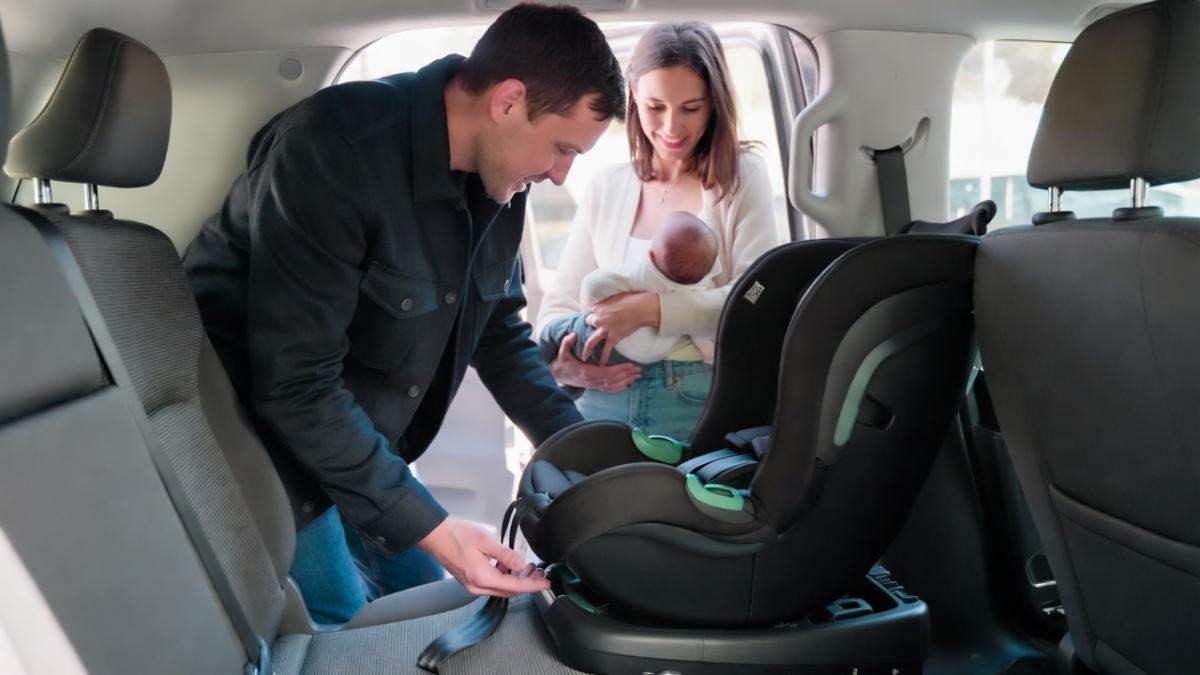 A young couple carefully installing a rear-facing infant car seat in the back of their SUV. The father kneels on the seat adjusting the base while the mother stands beside him holding their newborn baby wrapped in a blanket, both focused and smiling gently during the process.