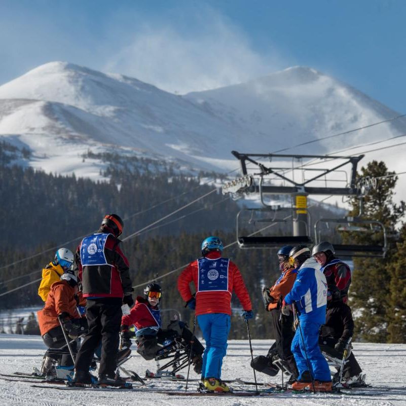 PSIA-AASI Continuing education course being conducted on snow, with standing and sitting skiers gathered in a group