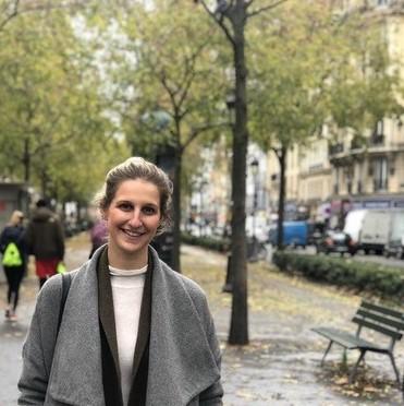 Photo of Krista Rappoccio, wearing a grey winter coat and standing on a busy city street sidewalk.
