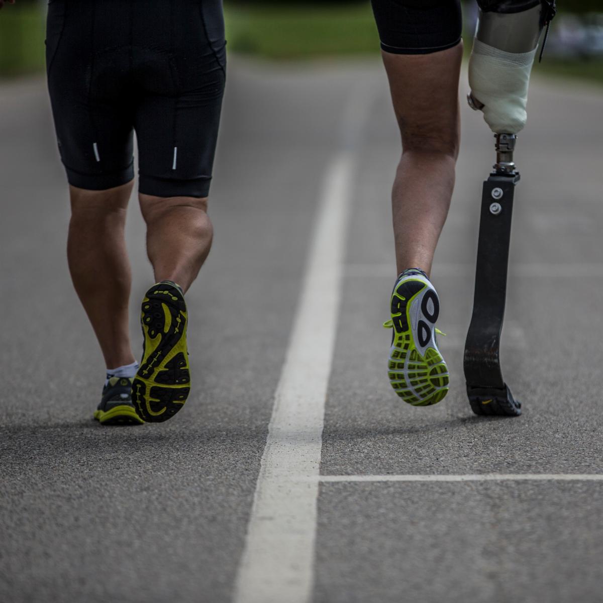 Two runners on a track. Runner on right has above knee amputation and is running with a blade prosthetic.