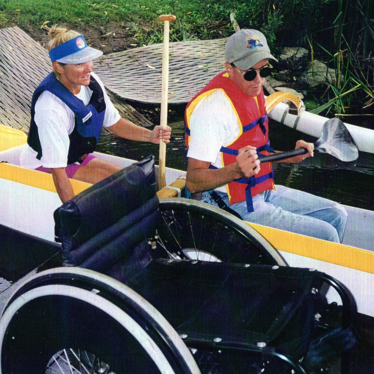 Man and woman seated in outrigger canoe with empty wheelchair in the foreground