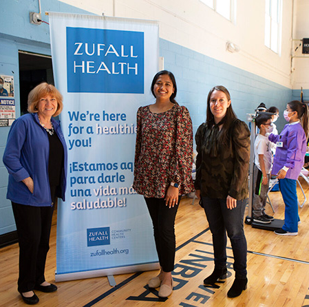 Three women including Eva Turbiner on far left stand in front of a Zufall roll-up banner in a gymnasium where a child's height is being measured by a medical professional