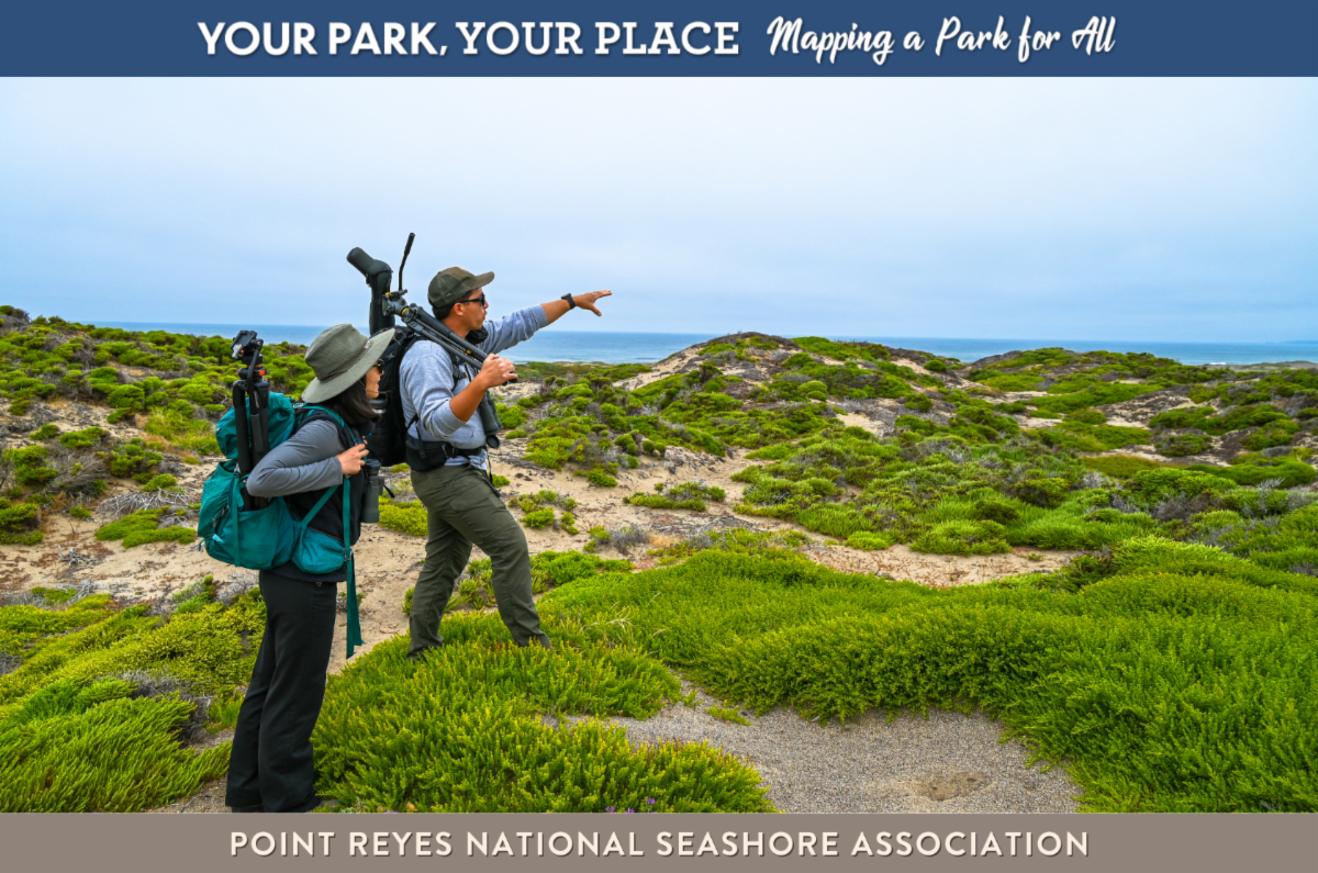 Two people stand on dunes covered in plants, with a blue-grey sky and water in the background. They both have backpacks on and are looking in the direction of where one of the people is pointing. He also has a camera on his shoulder.