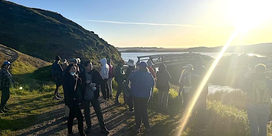 A group of people gathered on a trail with green hills water and a bridge in the background. The sun is in the top right corner of the image and a bright glare shines across the photo.