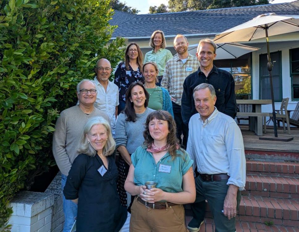 A group of eleven people standing outside on a staircase with a deck and hedge in the background.