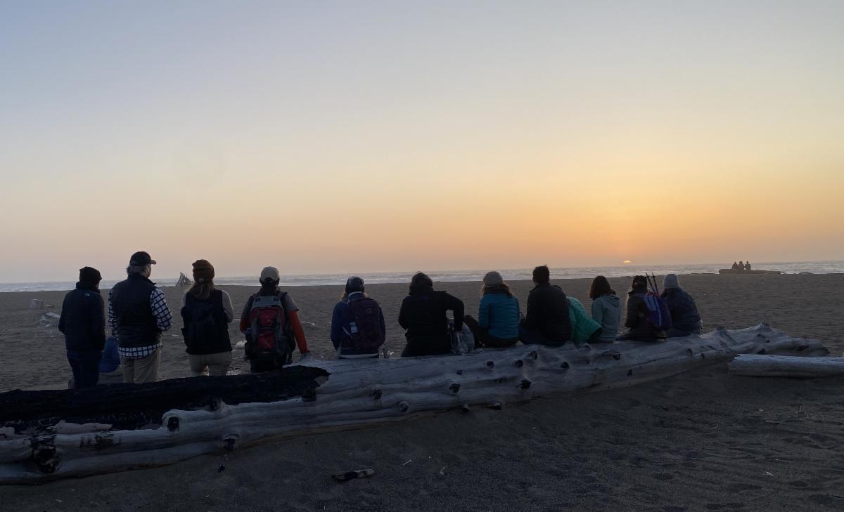 A group of people standing on the beach, looking out toward the setting sun.