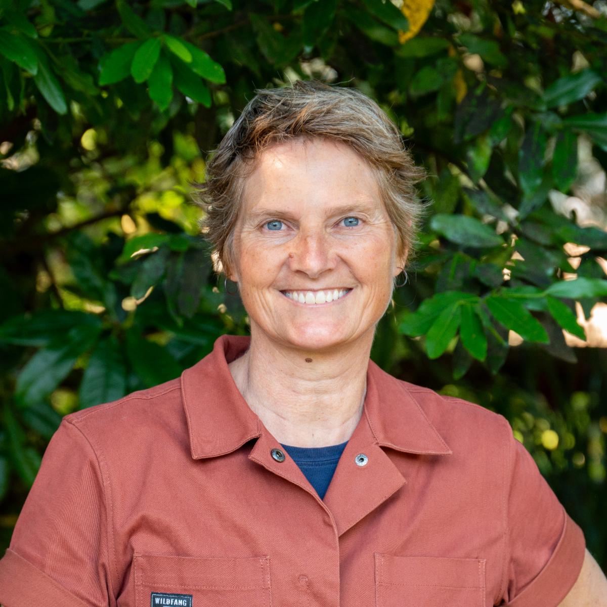 Biret wears a red jumpsuit and smiles in front of a green leafy hedge 