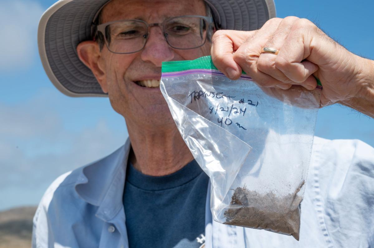 A man wearing blue clothing smiles as he holds up a transparent plastic bag containing a soil sample.