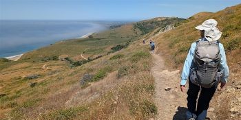  The back of a person hiking on a trail overlooking a coastline.