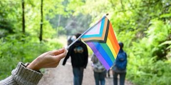 A hand holds a progress pride flag in front of a hiking trail.
