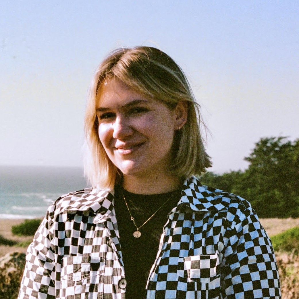 Emily is smiling at the camera, wearing a checkered jumpsuit. The ocean is visible in the background.