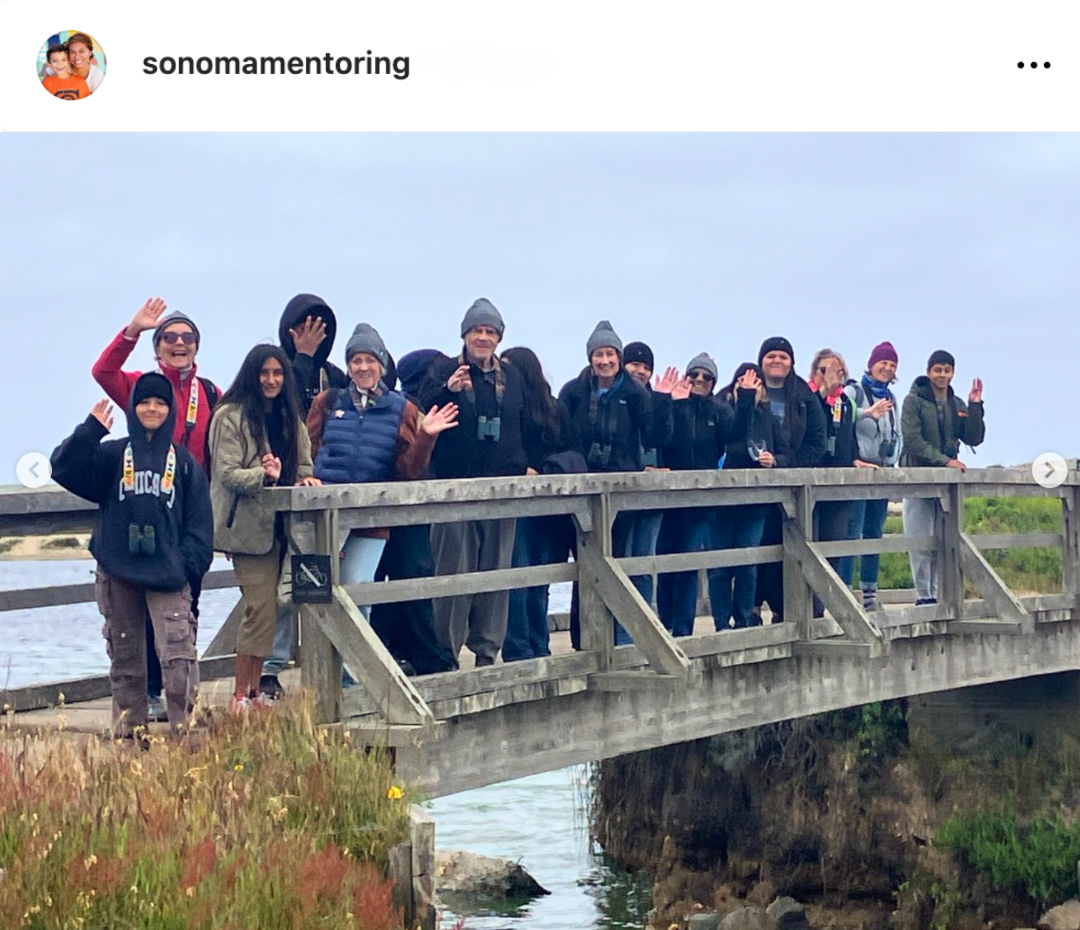 A group of people of all ages dressed for a chilly day stand on a bridge waving at the camera.
