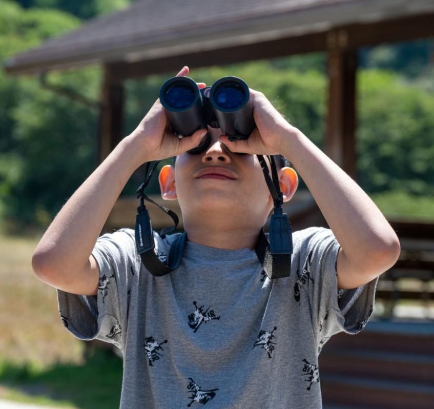 A child wearing a grey shirt looking through black binoculars
