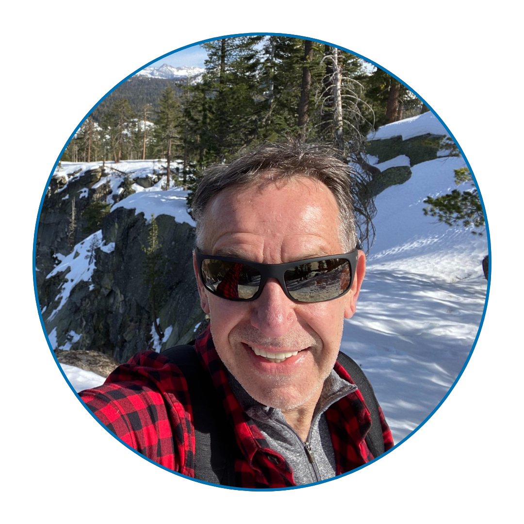 Stef smiling with a snowy landscape in the background. He wears sunglasses and a plaid shirt.