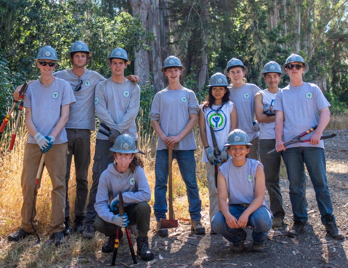 A group of young people standing together outdoors with grey shirts and hardhats.