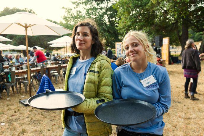 Two people wearing light blue shirts and holding black trays smile outdoors with a party visible in the background.