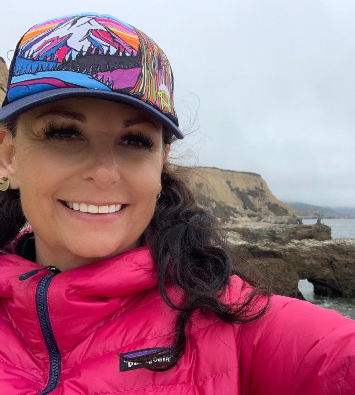 A woman in a hot pink jacket and baseball cap smiles at the camera with the ocean in the background.
