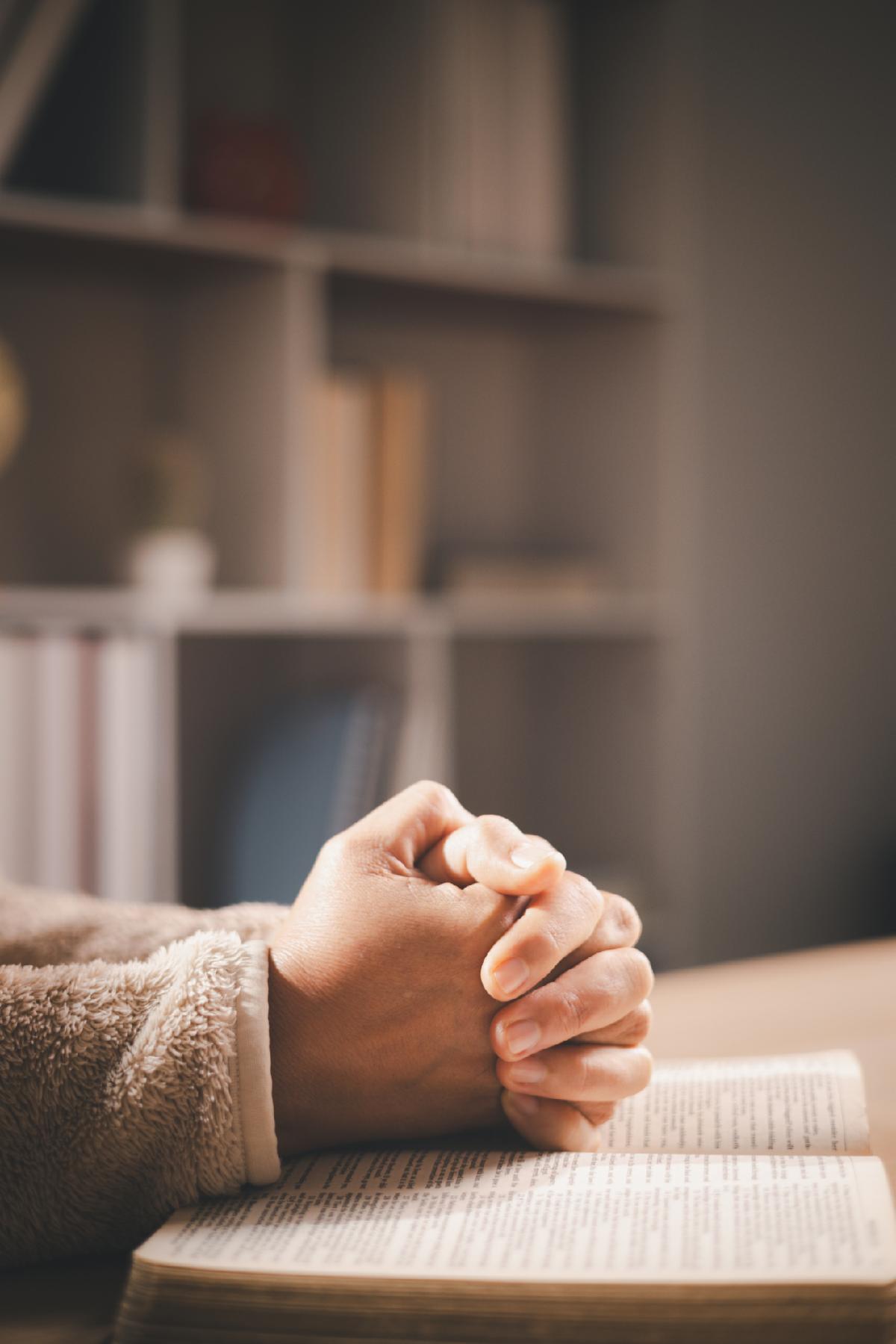 Clasped hands in prayer over an open book warm light and shallow depth of field symbolizing faith devotion reflection and hope for spiritual guidance during quiet study at home.