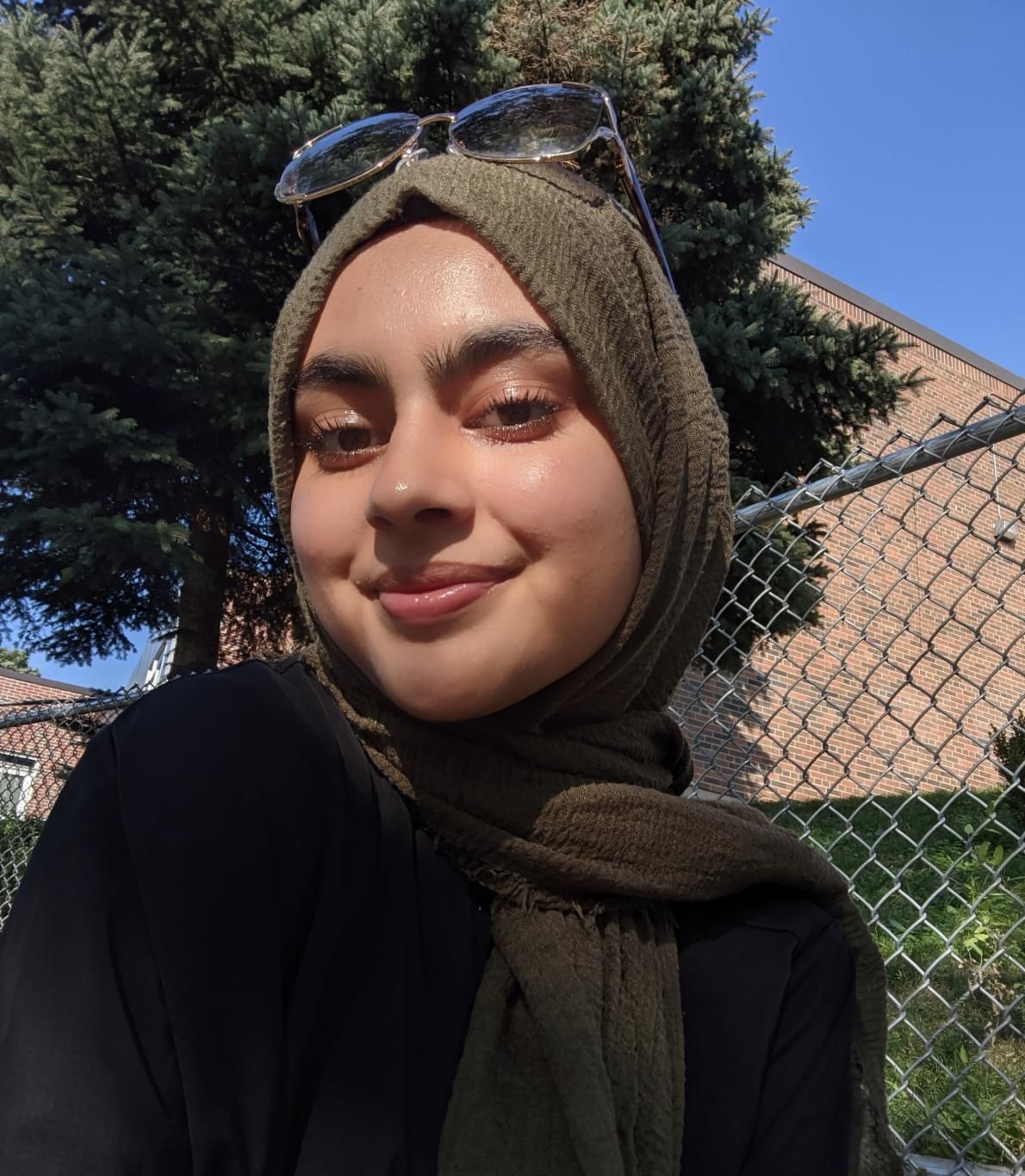 a brown appearing person wearing a light brown head scarf smiles with their head turned slightly to the side and gazing at the viewer in an outdoor setting with a leafy tree in the background