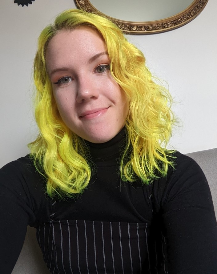 female presenting white person with shoulder length bright yellow wavy hair smiling slightly gazing at the viewer wearing a long sleeved black mock neck shirt