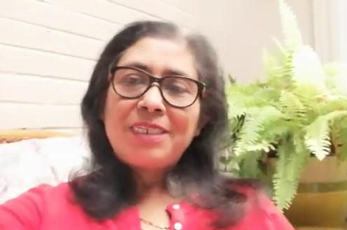 A brown person with long dark hair and glasses wearing a red shirt smiles at the viewer sitting next to a leafy plant