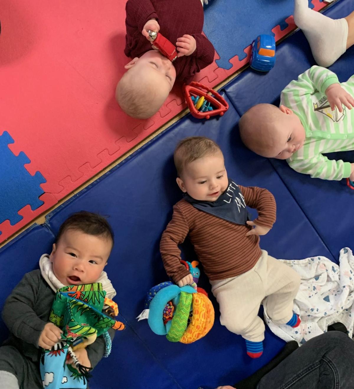 four babies lay on their backs on a mat with infant toys around them