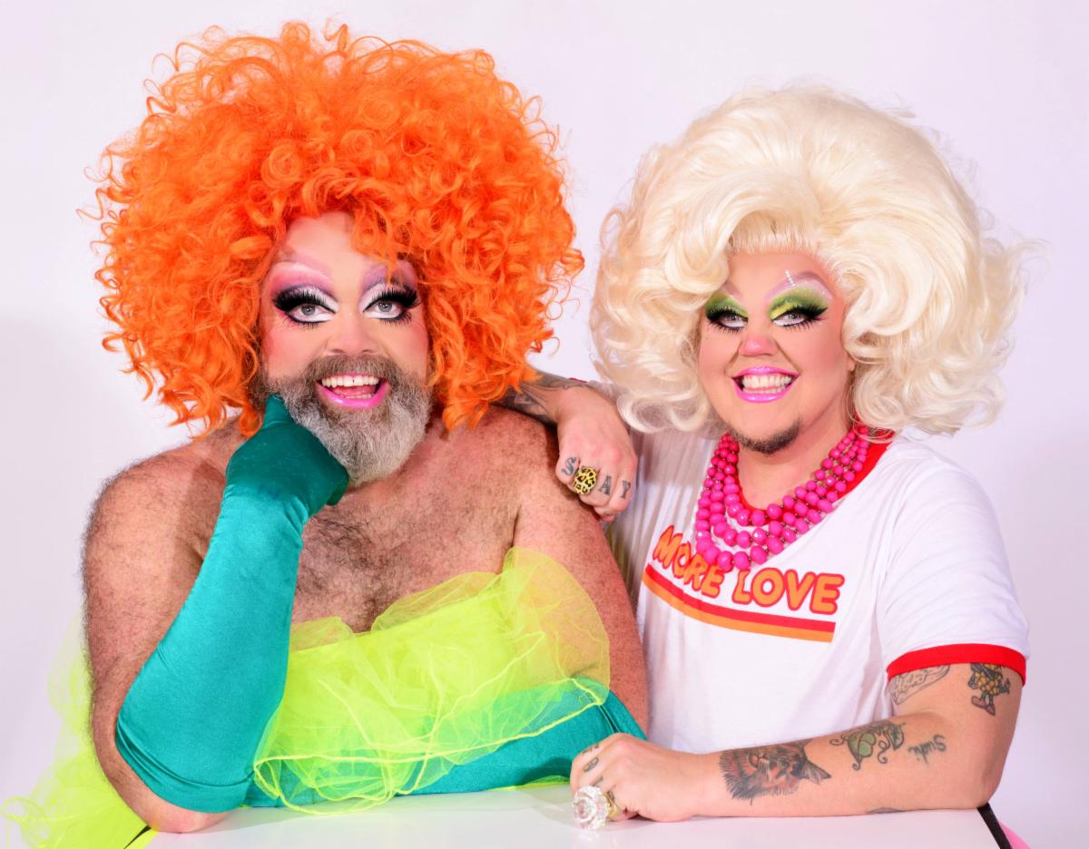 a bearded drag queen with big orange hair wearing an elegant chiffon dress and long turquoise gloves sits next to a drag queen with tattoos and big white hair wearing a white t shirt and pink beads